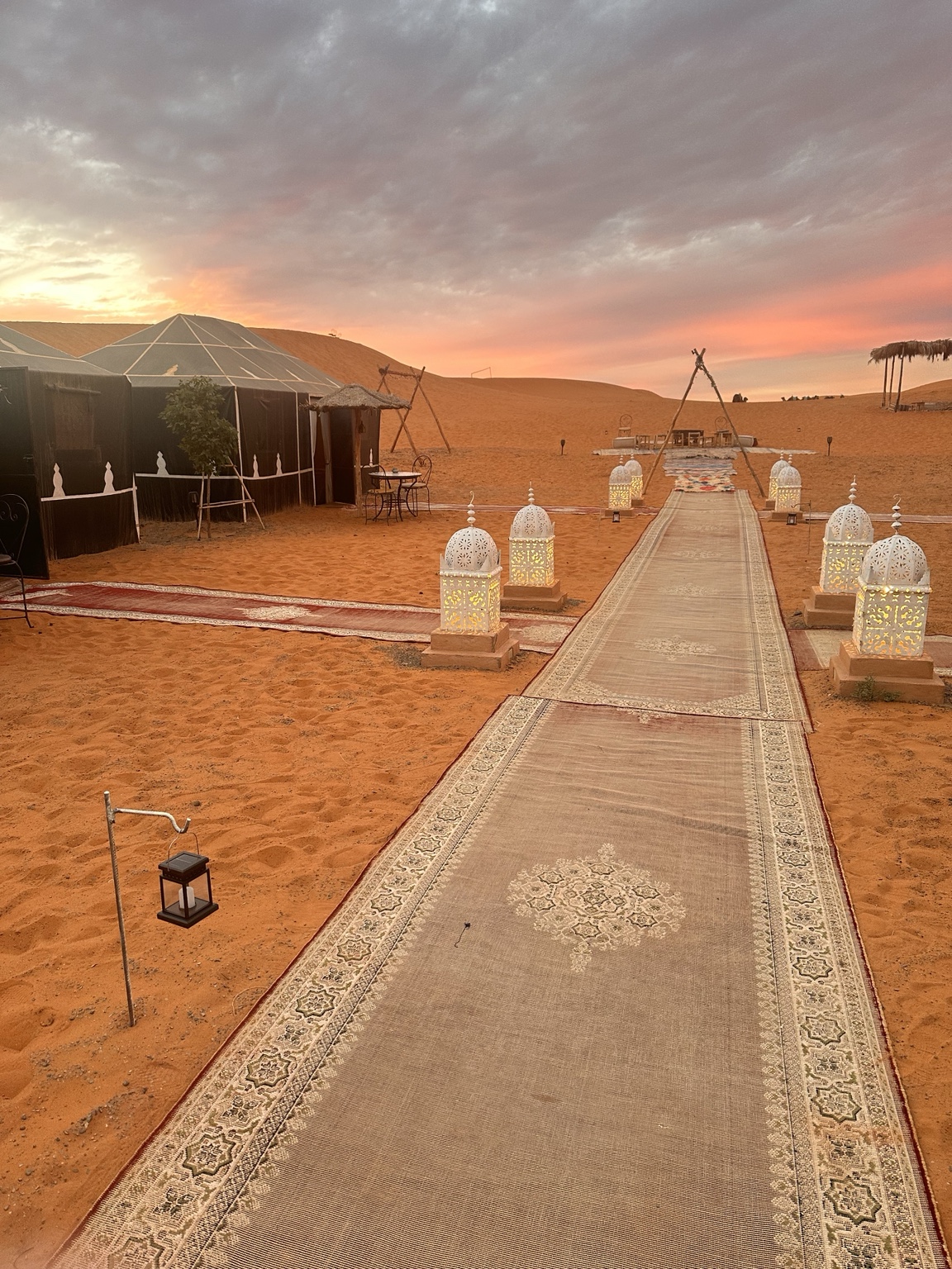 Lantern-lit path and tents at a Sahara camp under a pink sunset