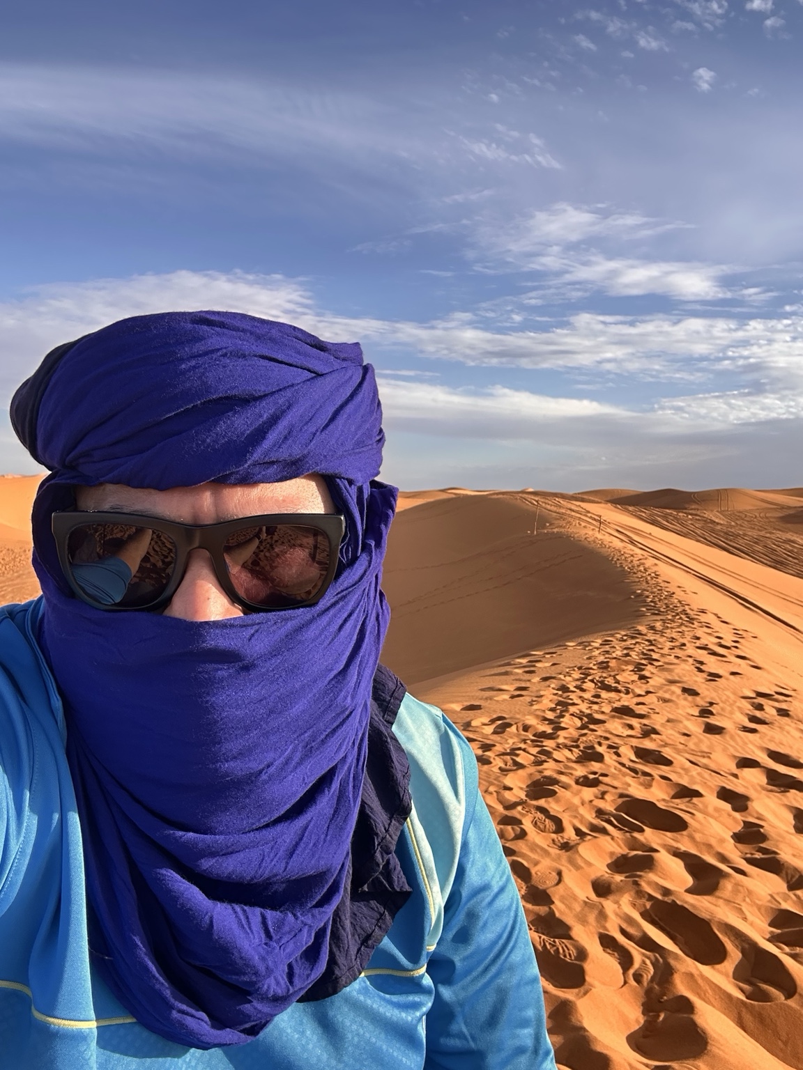 Gagan wearing a blue desert head wrap and sunglasses beside the Sahara dunes
